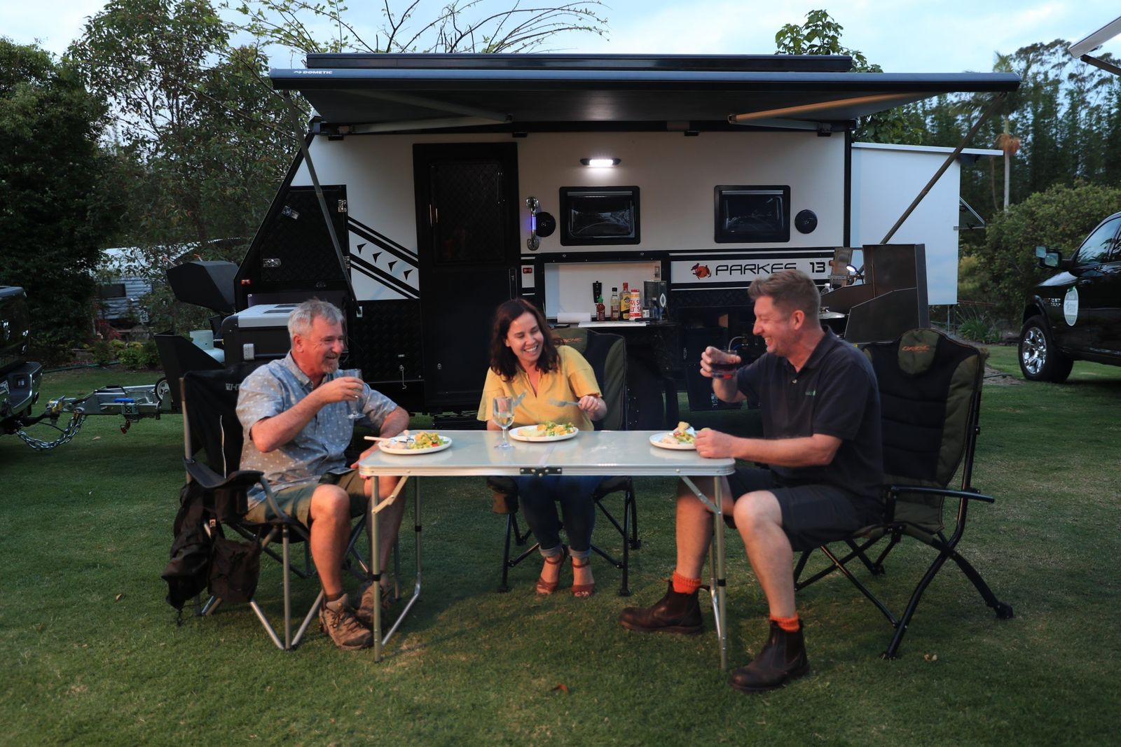 Three friends eating dinner under a caravan awning at dusk