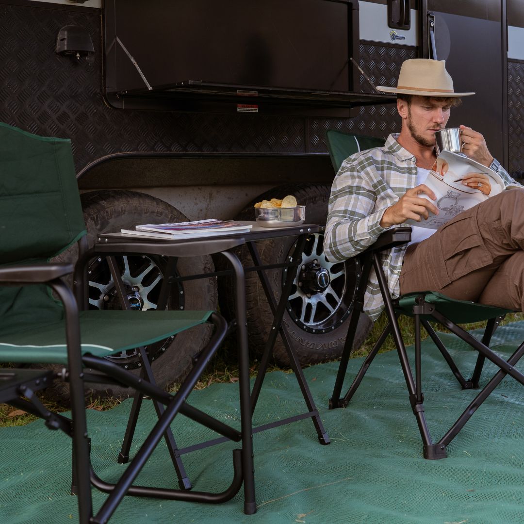 A man reading in a camp chair beside a caravan with a folding table