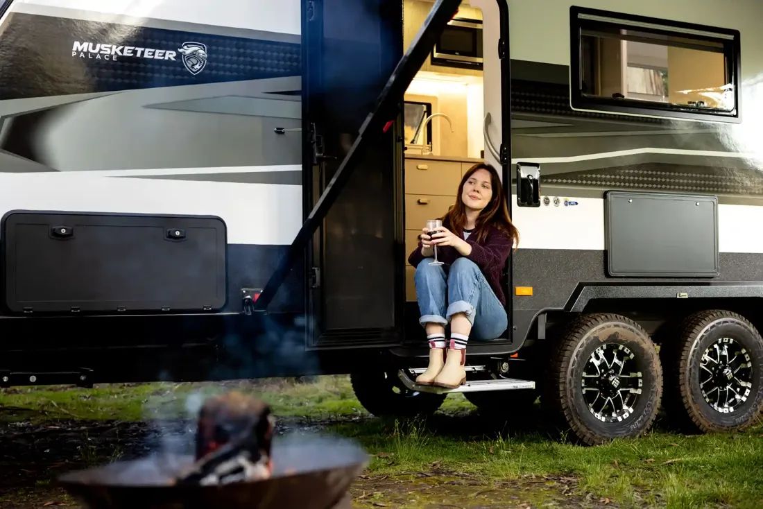 A woman sitting on the step of a caravan with a glass of wine by the campfire